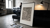 Death with skeleton takes a child print in a slim black frame leaning on a dark sofa in a concrete print studio