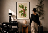 Holly Christmas Greetings print in a black frame on a sunlit plaster wall, a person looking on beside a mushroom lamp