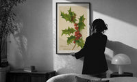 Holly Christmas Greetings print in a slim black frame being hung on a sunlit wall in a monochrome room