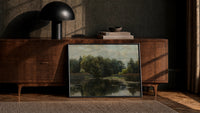 moody wooded pond/river landscape vintage art print print in a black frame leaning against a walnut console in lamplight