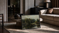 moody wooded pond/river landscape vintage art print print in a slim black frame leaning on a travertine coffee table