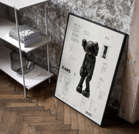 KAWS Companion Passing Through framed poster poster in a black frame leaning beside a shelf in a raw-plaster loft