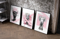 Pink KAWS Companion set of three black-framed prints leaning against a raw-plaster wall
