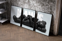 KAWS Reclining Companion set of three black-framed prints leaning against a raw-plaster wall
