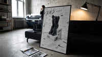 KAWS Companion Nike Swoosh Sketch framed poster print in a slim black frame leaning on a dark sofa in a concrete print studio