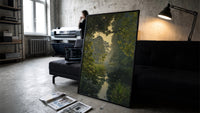 forest pond framed by overhanging branches vintage art print print in a slim black frame leaning on a dark sofa in a…