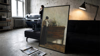 woman reading at a dresser in a quiet interior vintage art print print in a slim black frame leaning on a dark sofa in a…