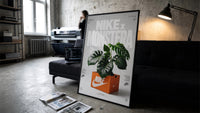 Nike Monstera Sneaker Box Green framed poster print in a slim black frame leaning on a dark sofa in a concrete print studio