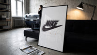 Nike Just Do It Swoosh framed poster print in a slim black frame leaning on a dark sofa in a concrete print studio