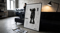 KAWS Companion Passing Through framed poster print in a slim black frame leaning on a dark sofa in a concrete print studio