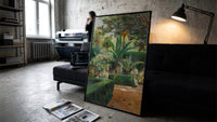 garden with palm trees and figures vintage art print print in a slim black frame leaning on a dark sofa in a concrete…