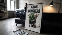 KAWS Companion Monstera Nike framed poster print in a slim black frame leaning on a dark sofa in a concrete print studio