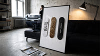 Saint Laurent Cest La Vie Skateboards framed poster print in a slim black frame leaning on a dark sofa in a concrete…