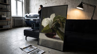white peony in dark glass vase vintage art print print in a slim black frame leaning on a dark sofa in a concrete print…