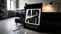 Nike Air History framed poster print in a slim black frame leaning on a dark sofa in a concrete print studio