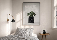 Nike x Monstera Garbage Bag print in a black frame above a bed with striped linens, a brass wall lamp and a sunlit window
