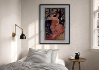 Mucha JOB print in a black frame above a bed with striped linens, a brass wall lamp and a sunlit window