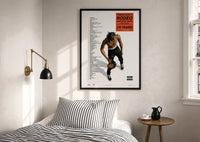 Travis Scott Rodeo Anniversary print in a black frame above a bed with striped linens, a brass wall lamp and a sunlit window