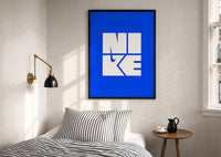 Nike Blue Typographic Logo print in a black frame above a bed with striped linens, a brass wall lamp and a sunlit window