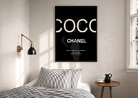 Coco Chanel Beauty Quote print in a black frame above a bed with striped linens, a brass wall lamp and a sunlit window