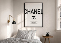 Coco Chanel Quote Typography print in a black frame above a bed with striped linens, a brass wall lamp and a sunlit window