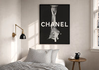 Chanel Black & White Luxury print in a black frame above a bed with striped linens, a brass wall lamp and a sunlit window