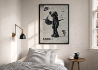 KAWS Companion Nike Swoosh print in a black frame above a bed with striped linens, a brass wall lamp and a sunlit window