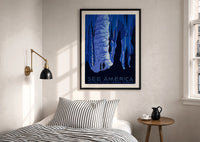 WPA See America Caverns print in a black frame above a bed with striped linens, a brass wall lamp and a sunlit window