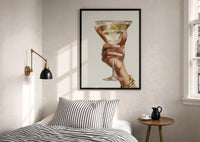 Martini Glass in Hand print in a black frame above a bed with striped linens, a brass wall lamp and a sunlit window