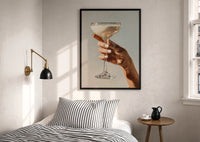 Cream Coupe in Hand print in a black frame above a bed with striped linens, a brass wall lamp and a sunlit window