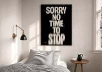 Sorry No Time To Stop print in a black frame above a bed with striped linens, a brass wall lamp and a sunlit window