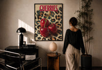 Cherries on Leopard print in a black frame on a sunlit plaster wall, a woman looking on beside a mushroom lamp
