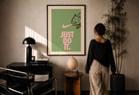 Just Do It Chameleon Green Pink print in a black frame on a sunlit plaster wall, a person looking on beside a mushroom lamp