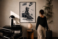 Nike x KAWS Air Force 1 print in a black frame on a sunlit plaster wall, a person looking on beside a mushroom lamp