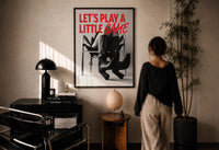 Let's Play a Little Game print in a black frame on a sunlit plaster wall, a person looking on beside a mushroom lamp
