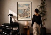 Kuniyoshi Water Scene with Boat print in a black frame on a sunlit plaster wall, a woman looking on beside a mushroom lamp