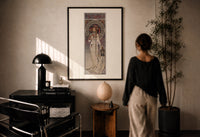 Mucha La Trappistine print in a black frame on a sunlit plaster wall, a woman looking on beside a mushroom lamp