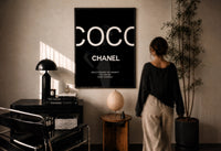Coco Chanel Beauty Quote print in a black frame on a sunlit plaster wall, a person looking on beside a mushroom lamp