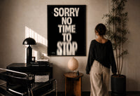 Sorry No Time To Stop print in a black frame on a sunlit plaster wall, a woman looking on beside a mushroom lamp