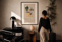Best Wishes Holly and Swallow print in a black frame on a sunlit plaster wall, a woman looking on beside a mushroom lamp