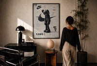 KAWS Companion Nike Swoosh print in a black frame on a sunlit plaster wall, a person looking on beside a mushroom lamp