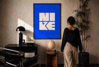 Nike Blue Typographic Logo print in a black frame on a sunlit plaster wall, a person looking on beside a mushroom lamp