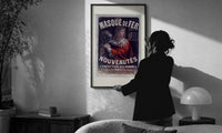 Cheret Au Masque de Fer print in a slim black frame being hung on a sunlit wall in a monochrome room