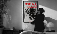 Let's Play a Little Game print in a slim black frame being hung on a sunlit wall in a monochrome room