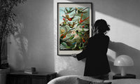 Haeckel Hummingbirds print in a slim black frame being hung on a sunlit wall in a monochrome room