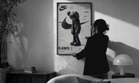 KAWS Companion Nike Swoosh print in a slim black frame being hung on a sunlit wall in a monochrome room