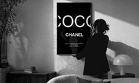 Coco Chanel Beauty Quote print in a slim black frame being hung on a sunlit wall in a monochrome room