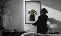 Nike x Monstera Garbage Bag print in a slim black frame being hung on a sunlit wall in a monochrome room