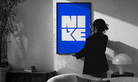 Nike Blue Typographic Logo print in a slim black frame being hung on a sunlit wall in a monochrome room