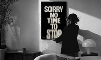 Sorry No Time To Stop print in a slim black frame being hung on a sunlit wall in a monochrome room